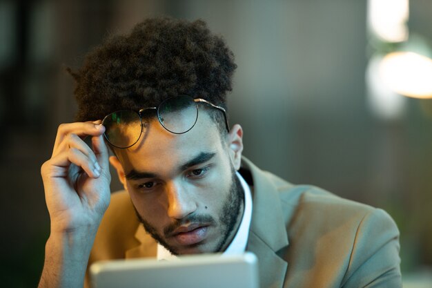 Ocupado jovem gerente negro com penteado afro tirando os óculos enquanto lê o artigo no tablet, ele está trabalhando até tarde