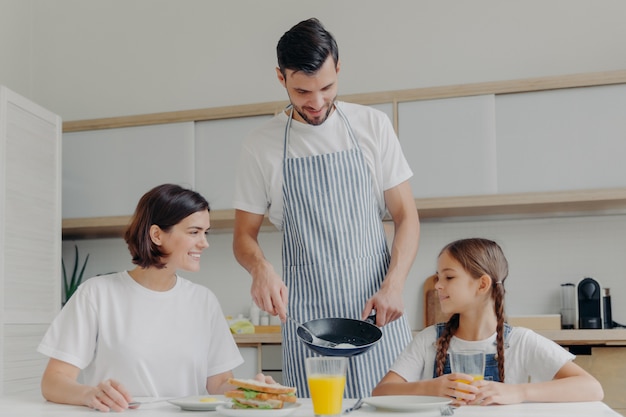 Ocupado padre cariñoso prepara un delicioso desayuno para esposa e hija, usa delantal, pone huevos fritos en un plato.