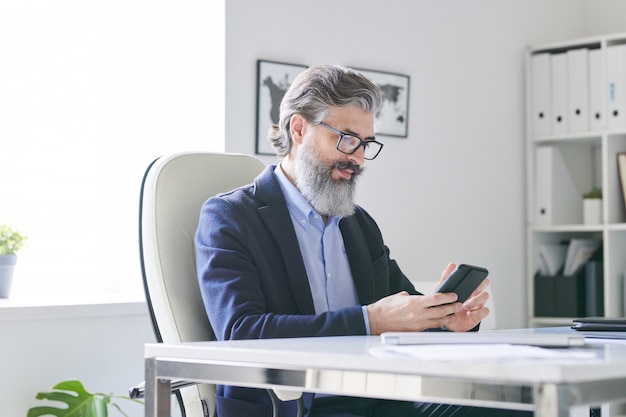 Ocupado profesional senior con cabello gris y barba desplazándose en el teléfono inteligente mientras verifica el horario de sus clientes o solicitantes en la oficina