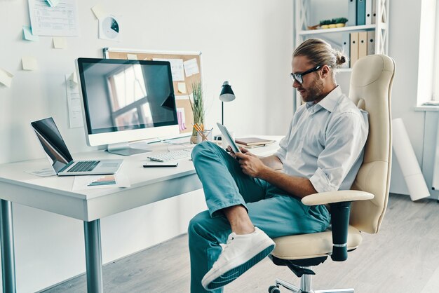 Ocupado trabajando. Apuesto joven en camisa con tableta digital mientras está sentado en la oficina