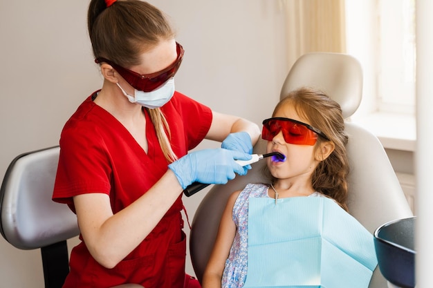 Odontología infantil Iluminación ultravioleta del procedimiento de llenado de dientes con fotopolímero Dentista infantil con gafas protectoras rojas trata y elimina la caries en un paciente