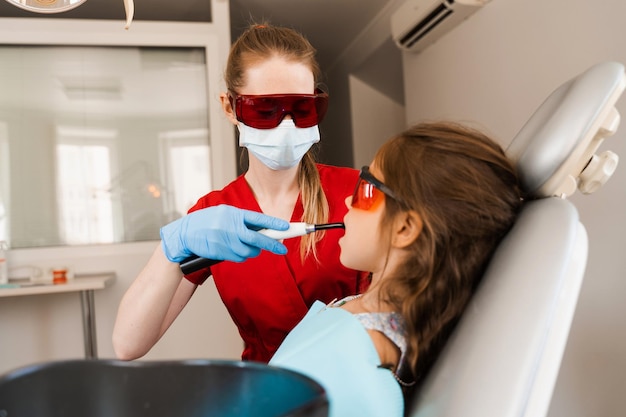 Odontología infantil Iluminación ultravioleta del procedimiento de llenado de dientes con fotopolímero Dentista infantil con gafas protectoras rojas trata y elimina la caries en un paciente