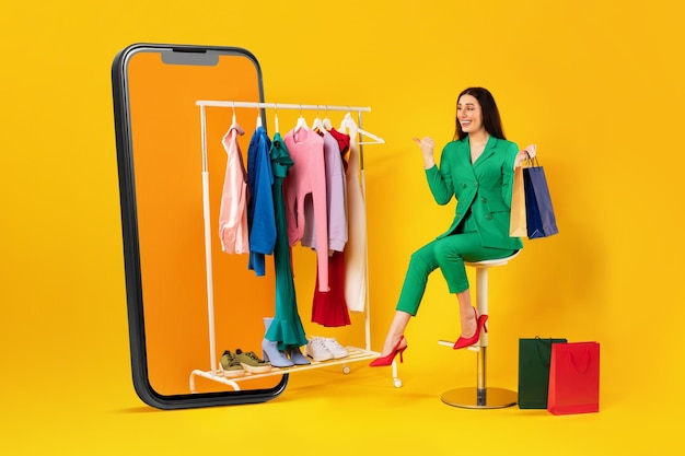 Oferta de compras móviles mujer adicta a las compras apuntando a la ropa en el riel de la ropa en un enorme teléfono celular sobre