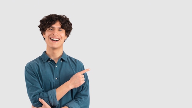 Oferta legal. Retrato de um cara feliz apontando de lado para o espaço da cópia contra a parede branca do estúdio. Jovem positivo oferecendo lugar para anúncio ou texto promocional e sorrindo para a câmera. Bandeira panorâmica