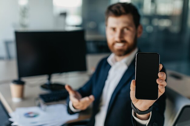 Oferta móvil Hombre de negocios que muestra un teléfono inteligente en blanco mientras está sentado en el escritorio en la maqueta de enfoque selectivo interior de la oficina