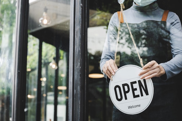 Offen. barista, kellnerin mit schutzgesichtsmaske, die offenes schild an der glastür im modernen café-café, café-restaurant, einzelhandelsgeschäft, kleinunternehmer, essen- und getränkekonzept aufdreht