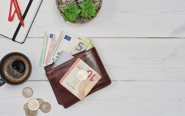 Offene Brieftasche für Herren aus Leder mit Euro-Banknoten-Münzen und Kreditkarte auf einem weißen Holztisch-Layout mit Kopierraum Planung von Einkäufen und Ausgaben eine Tasse Kaffee und ein Notizblock neben dem Tisch