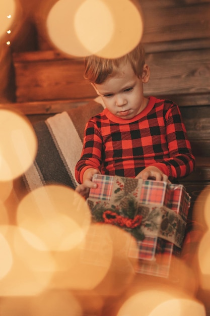 Offener, authentischer, glücklicher kleiner Junge im rot karierten Pyjama, der mit Geschenken in der weihnachtlich dekorierten Lodge sitzt