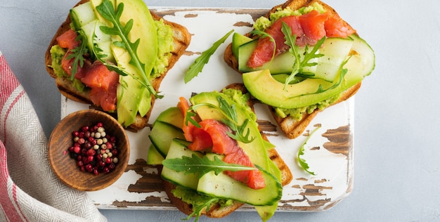 Offener Toast mit Forelle, Lachs, Avocado, Gurke und Rucola auf Holzständer auf grauem Betonhintergrund. Selektiver Fokus. Ansicht von oben. Platz kopieren