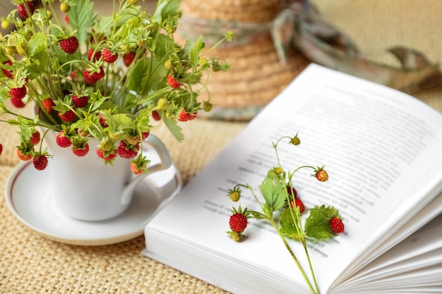 Offenes Buch, das in der Nähe eines sommerlichen Bündels wilder Erdbeerzweige mit roten Beeren liegt, die in einen Teebecher gelegt werden