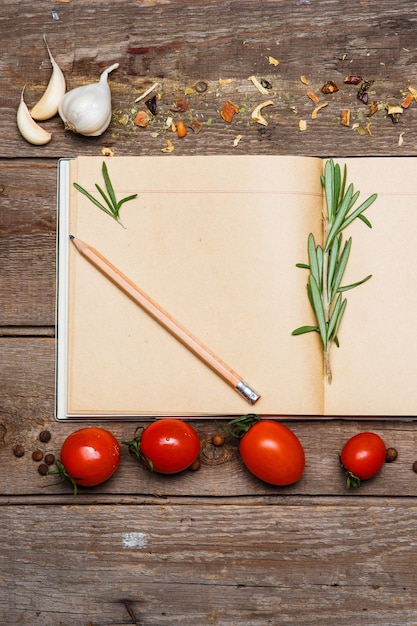 Offenes leeres Rezeptbuch auf braunem Holzhintergrund mit Tomaten, Knoblauch, Frühlingszwiebeln