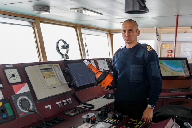 Foto offizier auf wache mit epirb auf der navigationsbrücke kaukasischer mann in blauem uniformpullover in notfallposition, der das funkleuchtfeuer auf der brücke des frachtschiffs anzeigt