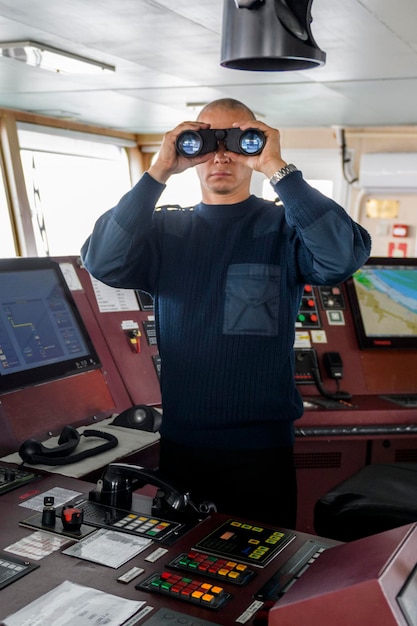 Oficial de plantão com binóculos na ponte de navegação Homem caucasiano de suéter uniforme azul na ponte do cargueiro