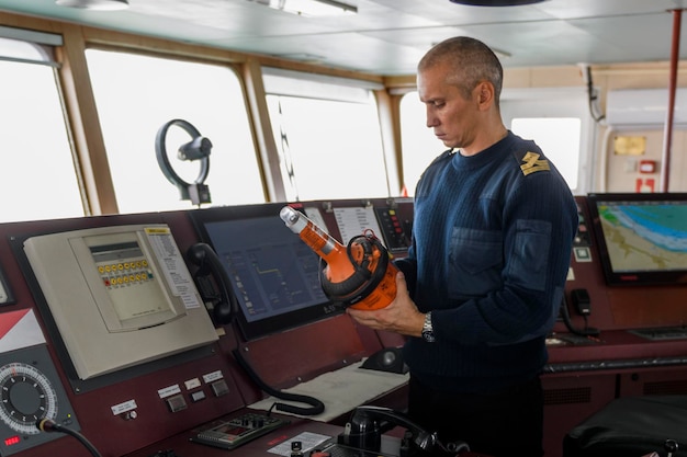 Oficial de serviço com EPIRB na ponte de navegação Homem caucasiano com suéter uniforme azul usando posição de emergência indicando farol de rádio na ponte do navio de carga
