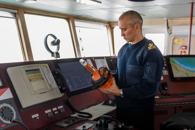 Oficial de guardia con EPIRB en el puente de navegación Hombre caucásico con suéter de uniforme azul que usa la posición de emergencia que indica la baliza de radio en el puente del buque de carga