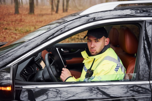 Oficial de policía masculino en uniforme verde sentado en un automóvil y trabajando con documentos.