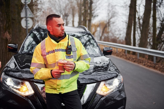 Oficial de policía masculino en uniforme verde tomando un descanso con donut en la carretera.