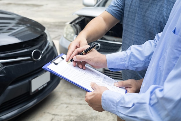 Oficial de seguros escribiendo en el portapapeles mientras el agente de seguros examina un auto negro después de un accidentexA