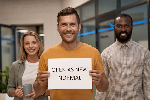 Oficina de apertura joven trabajador de oficina feliz mostrando papel con texto abierto como nuevo normal en la cámara