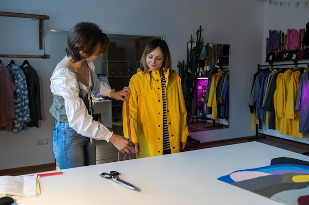 Oficina de designer de moda com alfaiate feminina medindo e ajustando jaqueta de chuva amarela sob medida