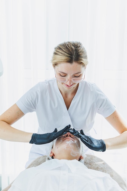 Oficina de un dermatólogo o cosmetólogo que trabaja con pacientes femeninas sentadas en una mesa de masaje masajeando el rostro de una mujer joven y haciendo otros procedimientos para la piel facial Cuidado de la piel facial