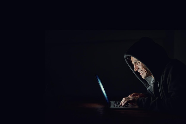 Oficina oscura y hombre en portátil para piratear programación en línea y ciberseguridad por la noche Desarrollador de software o hacker con capucha codificando contraseña para firewall o acceso a la red en fondo negro