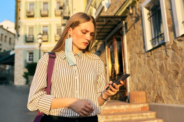 Oficinista de mujer de negocios joven en máscara protectora médica con smartphone en manos, máscara en la cara abierta del oído. Mujer en la calle de la ciudad. Estilo de vida, negocios en una epidemia, pandemia, mala ecología