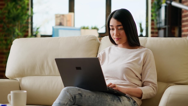 Oficinista sonriente en casa trabajando remotamente en una laptop mientras se sienta en un sofá en la sala de estar. Persona adulta joven que hace trabajo remoto en una computadora portátil moderna mientras está en casa.