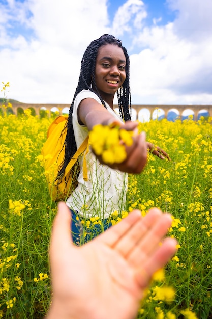 Ofreciendo una flor a una chica étnica negra con trenzas a un viajero en un campo de flores amarillas