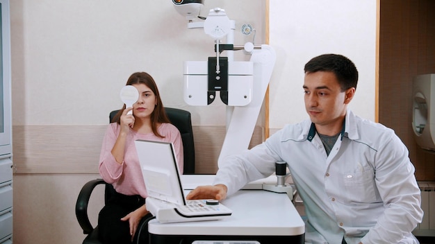 Oftalmología joven comprobando su agudeza visual cerrando un ojo y leyendo cartas de un médico sentado cerca