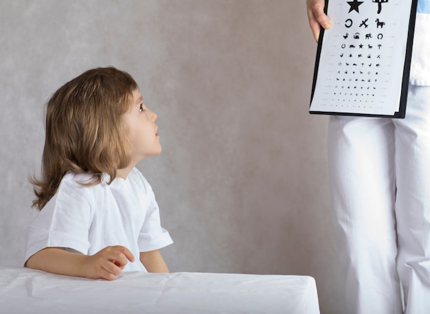 Foto oftalmologista verifica a visão de menino de 5 anos por meio de gráfico oftalmológico infantil.