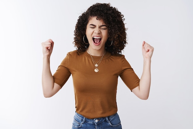 Foto oh sim. mulher animada, feliz e emotiva, cabelos cacheados escuros gritam bem alto felicidade alegria alcançar o sucesso bom negócio fechar olhos gritando sim cerrar os punhos triunfo torcer comemorar vitória ganhando na loteria