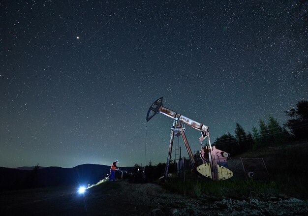 Foto oilman trabajando el turno de noche en el pozo de petróleo trabajador comprobando los parámetros de funcionamiento de la bomba de aceite jack en el fondo hermoso cielo estrellado y siluetas colinas de montaña