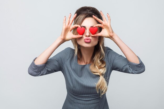 Ojos amorosos. Hermosa mujer joven caucásica romántica sosteniendo dos corazones de San Valentín frente a sus ojos como gafas mientras está de pie contra el fondo gris. Copie el espacio, interior, tiro del estudio, aislado