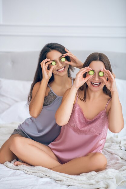 Ojos de pepino. Dos lindas chicas jóvenes con rodajas de pepino en las manos divirtiéndose y mirando feliz