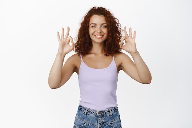 OK. Garota ruiva satisfeita mostra sinal de aprovação de OK, sorrindo satisfeito, recomendando, garantia de qualidade, loja de elogios, em pé no branco.