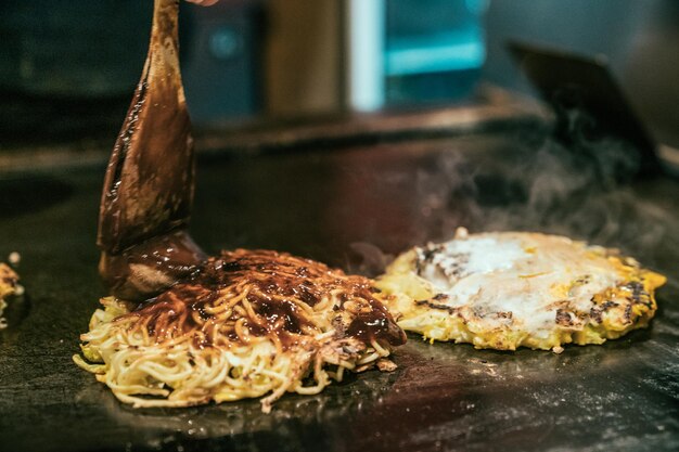 Okonomiyaki auf Teppanyaki-Pfanne. Der Küchenchef gießt salzige Sauce mit einem Pinsel auf Pfannkuchen nach japanischer Art, um ihn köstlich zu machen. Heißes leckeres traditionelles Straßenessen auf Metalleisenplatte, die mit Rauch kocht.