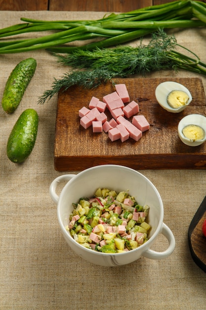 Okroshka em uma tigela de sopa em uma mesa de madeira ao lado de verdes em pranchas e uma faca. Foto vertical