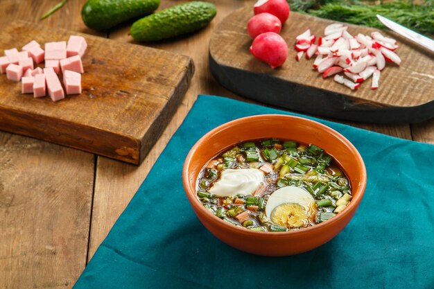 Okroshka en un plato de arcilla sobre la mesa sobre un mantel junto a verduras en tablas. Foto horizontal