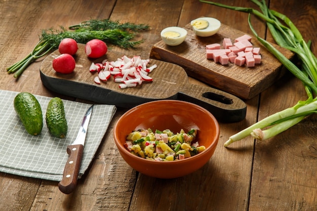 Okroshka en un plato de arcilla sobre una mesa sobre una mesa de madera junto a verduras en tablas y un cuchillo.