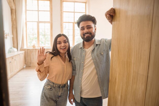 Foto olá, felizes cônjuges indianos conhecendo alguém perto da porta aberta, mulher acenando com a mão