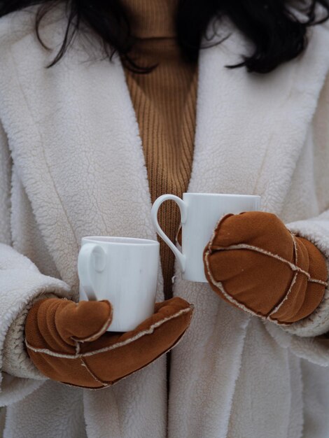 Olá inverno aconchegante, clima de inverno, uma garota com um casaco de pele branco e luvas quentes segura duas xícaras de chá dentro dela ...