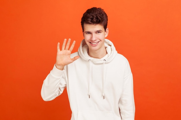 Olá jovem bonito em moletom casual elegante dizendo oi com sorriso no rosto feliz em vê-lo conhecido Tiro de estúdio interior isolado em fundo laranja