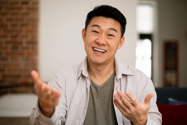 Olá Retrato de homem asiático maduro feliz gesticulando e falando para a câmera sentado no sofá em casa