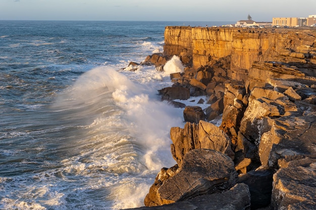 Las olas del mar chocan contra un acantilado rocoso en la hora dorada, la gente está de pie en la cima del acantilado y disfruta