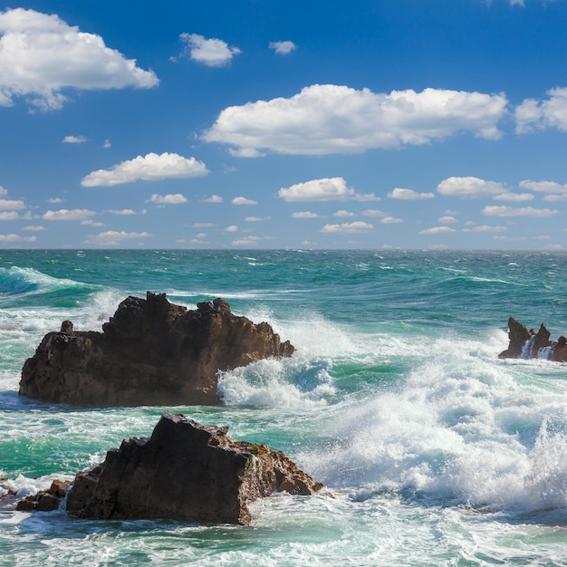 Olas turquesas del océano golpeando las rocas de la costa con cielo azul y nubes blancas en el fondo