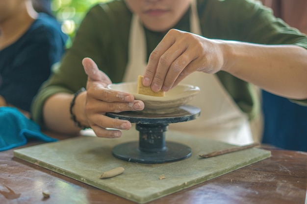 Oleiro de homem trabalhando na roda de oleiro fazendo pote de cerâmica de barro no conceito de arte de oficina de cerâmica Foco mão jovem anexando parte do produto de barro ao futuro produto de cerâmica Oficina de cerâmica