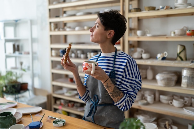 Oleiro feminino em avental sujo segurando caneca de barro artesanal com café desfrutando de almoço na oficina