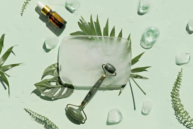 Óleo essencial de rolo facial de pedra de jade e creme artesanal com cubos de gelo e folhas de samambaia exóticas em fundo verde menta