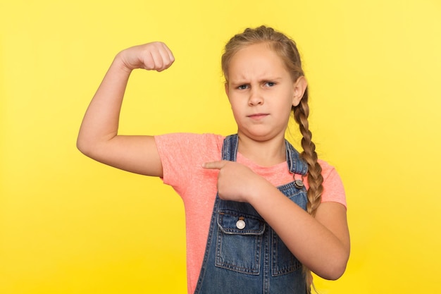 Olha, eu sou forte. Retrato de menina corajosa com trança em macacão jeans apontando para bíceps, criança mostrando poder e confiança para crescer saudável. tiro de estúdio interno isolado em fundo amarelo